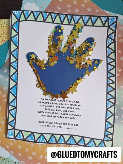 A blue paper handprint on white paper is decorated with gold glitter and star-shaped confetti. The paper has a geometric blue border and a printed poem about a child’s growing hands. @GLUEDTOMYCRAFTS is labeled at the bottom.