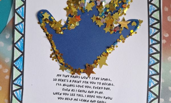 A blue paper handprint on white paper is decorated with gold glitter and star-shaped confetti. The paper has a geometric blue border and a printed poem about a child’s growing hands. @GLUEDTOMYCRAFTS is labeled at the bottom.