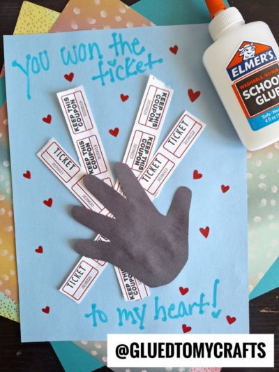 A handmade HANDPRINT FATHER'S DAY craft features a hand cutout on blue paper with paper tickets radiating from it, surrounded by red hearts. The text says, “you won the ticket to my heart!” Glue and colorful paper are scattered nearby.