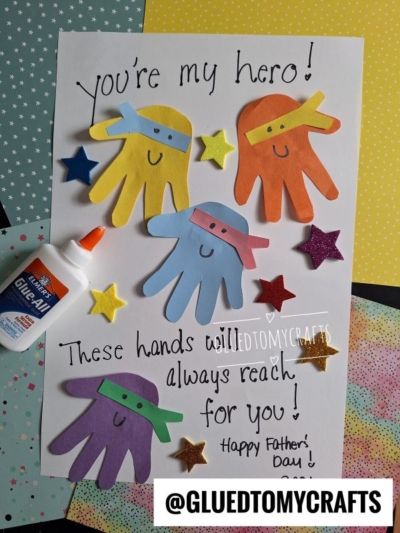A handmade HANDPRINT FATHER'S DAY card features colorful cut-out paper handprints as superheroes with masks, surrounded by foam stars and glue. The message reads, "You’re my hero! These hands will always reach for you! Happy Father’s Day!.