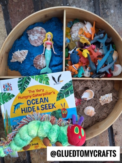 A wooden tray is divided into three sections with blue kinetic sand, sea animals, a mermaid, brown sand, shells, and toy sharks—perfect for kinetic sand play ideas. In front is The Very Hungry Caterpillar’s Ocean Hide & Seek book and a plush caterpillar.