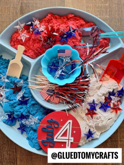 A round tray divided into sections holds red, blue, and tan colored sand, plastic stars, American flag toothpicks, scoops, and festive Fourth of July-themed decorations—perfect for patriotic kinetic sand play ideas. A July 4 sign and @GLUEDTOMYCRAFTS label are visible.