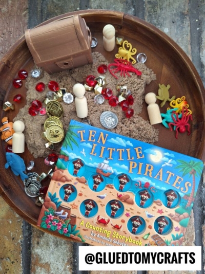 A wooden tray holds a pirate-themed counting book, toy coins, jewels, a treasure chest, miniature sea creatures, peg dolls, and kinetic sand—perfect for fun kinetic sand play ideas. The Instagram handle @GLUEDTOMYCRAFTS is at the bottom.