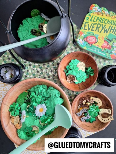 A St. Patrick’s Day craft setup with green sensory sand, gold coins, shamrock charms, daisies, a cauldron, the book Leprechauns Everywhere, and large spoons—perfect for festive kinetic sand play ideas on the table.