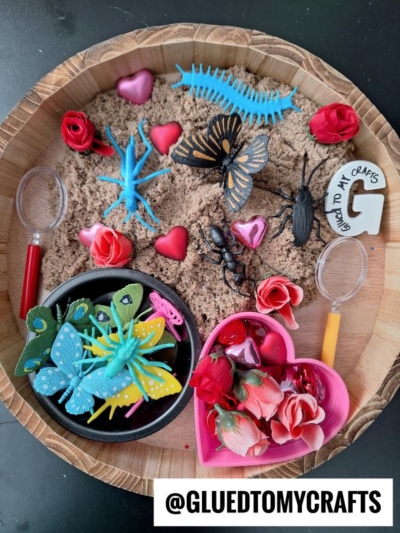 A round tray featuring kinetic sand play ideas with toy insects, red and pink artificial roses, heart objects, magnifying glasses, and play sand in sections. Includes a black bowl with colorful butterflies and a heart-shaped container of roses.