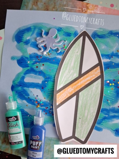 A handmade surfboard craft on blue painted paper with an octopus sticker, glitter, and two Tulip paint bottles captures a fun paint splat beach wave vibe. @GLUEDTOMYCRAFTS is written on the paper twice.