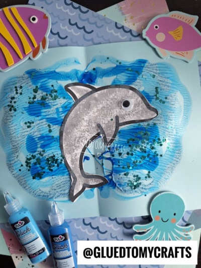 A handmade dolphin craft on blue paper, surrounded by sea-themed decorations like a fish and an octopus, with blue paint and glitter creating a water effect. Two bottles of blue paint are nearby. @GLUEDTOMYCRAFTS is on the image.