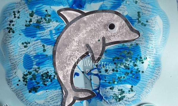 A handmade dolphin craft on blue paper, surrounded by sea-themed decorations like a fish and an octopus, with blue paint and glitter creating a water effect. Two bottles of blue paint are nearby. @GLUEDTOMYCRAFTS is on the image.