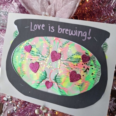 A handmade craft of a black cauldron with colorful green, yellow, and pink paint inside, decorated with glittery pink hearts. The text Love is brewing! is written above. Instagram handle @GLUEDTOMYCRAFTS is at the bottom.
