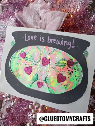 A handmade craft of a black cauldron with colorful green, yellow, and pink paint inside, decorated with glittery pink hearts. The text Love is brewing! is written above. Instagram handle @GLUEDTOMYCRAFTS is at the bottom.