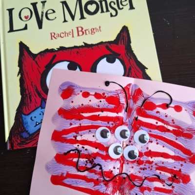 A children’s book titled Love Monster by Rachel Bright sits behind a playful paint splat love monster craft, made with paint and googly eyes on pink paper. The photo is labeled @GLUEDTOMYCRAFTS.