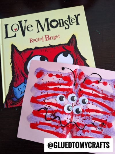 A children’s book titled Love Monster by Rachel Bright sits behind a playful paint splat love monster craft, made with paint and googly eyes on pink paper. The photo is labeled @GLUEDTOMYCRAFTS.