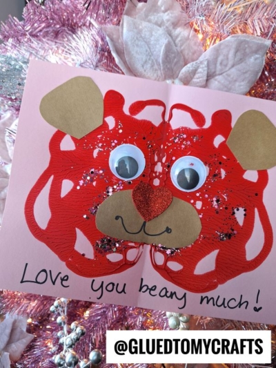 A handmade Valentines card featuring a red PAINT SPLAT CRAFTS design resembling a bear’s face, with googly eyes, a glittery red nose, and heart-shaped ears. Text reads "Love you beary much!" in black marker.