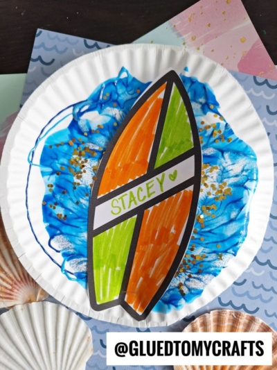 A paper plate craft with a blue and white paint splat background evokes a beach wave, accented by gold glitter and a colorful surfboard labeled Stacey in green, resting on beach-themed paper. @GLUETOMYCRAFTS appears at the bottom.