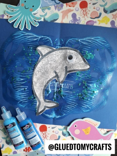 A handmade dolphin craft on blue paper, surrounded by ocean-themed stickers like an octopus and fish. Blue glitter and paint create a textured sea effect. Two blue paint bottles and a @GLUEDTOMYCRAFTS label are visible.