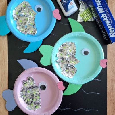 Three colorful paper plate fish crafts with foil scales, construction paper fins, and googly eyes are displayed on a blackboard, alongside glue, a roll of aluminum foil, and the @GLUEDTOMYCRAFTS label.