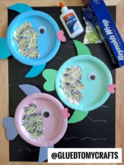 Three colorful paper plate fish crafts with foil scales, construction paper fins, and googly eyes are displayed on a blackboard, alongside glue, a roll of aluminum foil, and the @GLUEDTOMYCRAFTS label.