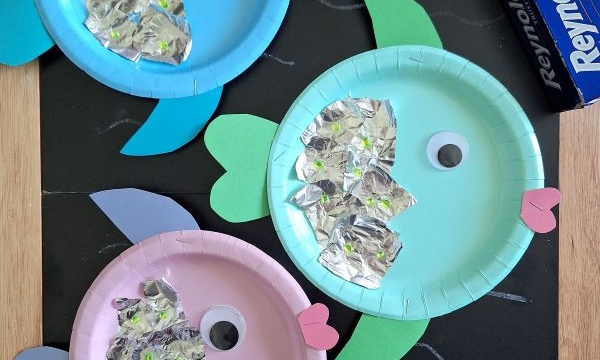 Three colorful paper plate fish crafts with foil scales, construction paper fins, and googly eyes are displayed on a blackboard, alongside glue, a roll of aluminum foil, and the @GLUEDTOMYCRAFTS label.