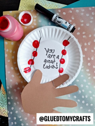 A HANDPRINT FATHER'S DAY craft: a paper plate with "You are a great catch!" written on it, decorated with red paint fingerprints like cherries and a brown paper hand attached, surrounded by craft supplies. @GLUEDTOMYCRAFTS at the bottom.