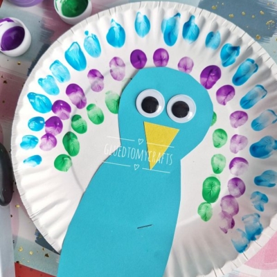 A paper plate decorated as a colorful peacock with blue, purple, and green painted fingerprints for feathers, a blue paper body, large googly eyes, and a yellow beak. @GLUETOMYCRAFTS watermark is visible.