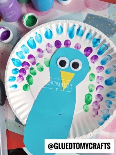 A paper plate decorated as a colorful peacock with blue, purple, and green painted fingerprints for feathers, a blue paper body, large googly eyes, and a yellow beak. @GLUETOMYCRAFTS watermark is visible.