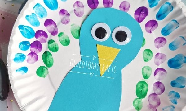 A paper plate decorated as a colorful peacock with blue, purple, and green painted fingerprints for feathers, a blue paper body, large googly eyes, and a yellow beak. @GLUETOMYCRAFTS watermark is visible.