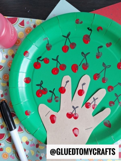 A green paper plate decorated with red fingerprint cherries and black marker stems. A tan paper cut-out of a hand is glued to the plate, also decorated with cherries. Arts and crafts materials surround the plate.