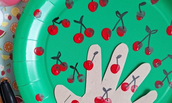 A green paper plate decorated with red fingerprint cherries and black marker stems. A tan paper cut-out of a hand is glued to the plate, also decorated with cherries. Arts and crafts materials surround the plate.