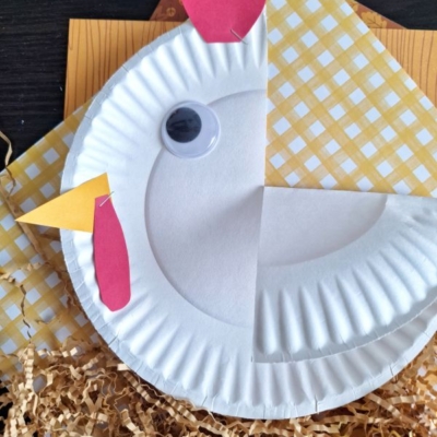A paper plate is decorated to look like a chicken with red and yellow paper for the beak, wattle, and comb, a large googly eye, and is placed on yellow paper and shredded brown paper.