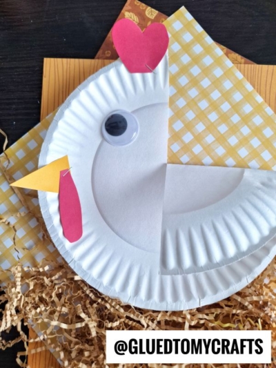 A paper plate is decorated to look like a chicken with red and yellow paper for the beak, wattle, and comb, a large googly eye, and is placed on yellow paper and shredded brown paper.
