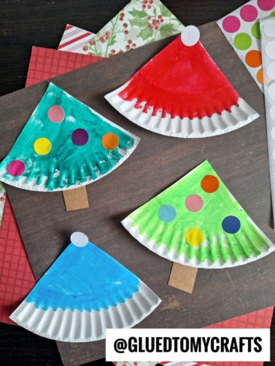 Four colorful paper plate Christmas tree crafts, each painted in different colors and decorated with round stickers, are displayed on a table with craft supplies and patterned paper in the background.