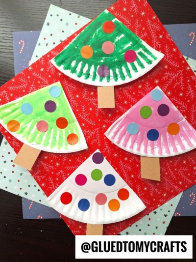 Four paper plate Christmas trees, decorated with colorful circle stickers and brown paper trunks, are displayed on festive red and blue holiday paper backgrounds. @GLUEDTOMYCRAFTS is written at the bottom.
