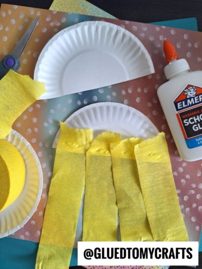 Paper plates, yellow crepe paper strips, scissors, and a bottle of Elmer’s school glue are arranged on colorful polka dot paper. The image also displays the text @GLUEDTOMYCRAFTS.