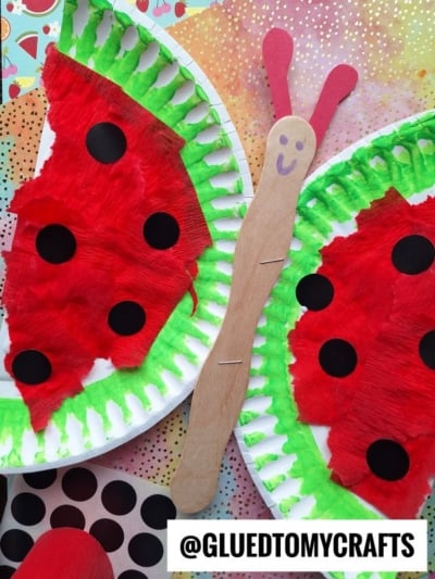 A colorful craft made from paper plates painted to look like watermelon slices, with a popsicle stick forming a caterpillar with a smiley face and red antennae. The background features patterned paper.