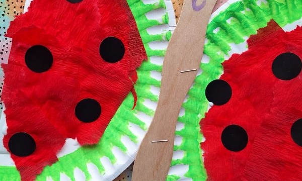 A colorful craft made from paper plates painted to look like watermelon slices, with a popsicle stick forming a caterpillar with a smiley face and red antennae. The background features patterned paper.