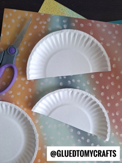 Two white paper plates cut in half are placed on a colorful, dotted background with a pair of purple-handled scissors nearby. The bottom of the image has the text @GLUEDTOMYCRAFTS.