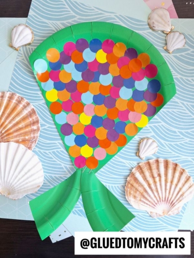 A colorful fish tail craft made from green paper and covered in round, multicolored paper scales, surrounded by seashells on a blue patterned background—perfect for coding label crafts and creative projects.