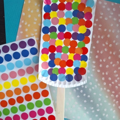 A craft project resembling a colorful ice cream popsicle made with multicolored dot stickers on a white paper shape, attached to a wooden stick, placed on pastel papers. A sticker sheet and @GLUEDTOMYCRAFTS text are also visible.
