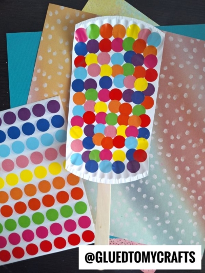 A craft project resembling a colorful ice cream popsicle made with multicolored dot stickers on a white paper shape, attached to a wooden stick, placed on pastel papers. A sticker sheet and @GLUEDTOMYCRAFTS text are also visible.
