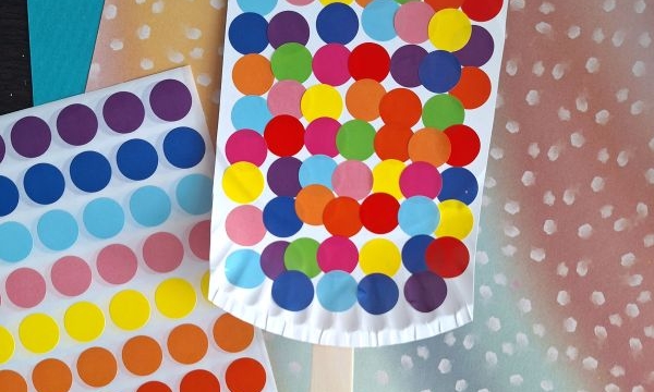 A craft project resembling a colorful ice cream popsicle made with multicolored dot stickers on a white paper shape, attached to a wooden stick, placed on pastel papers. A sticker sheet and @GLUEDTOMYCRAFTS text are also visible.