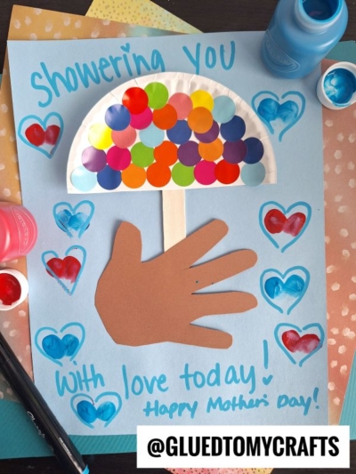 A handmade Mother’s Day card features a brown paper hand, a paper plate umbrella with colorful dots, painted hearts, and a coding label crafts touch with the words “Showering You With Love Today! Happy Mother’s Day!” on blue paper. @GluedToMyCrafts.