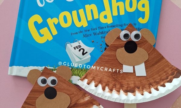 A childrens book titled How to Catch a Groundhog is displayed with two paper plate groundhog crafts, each featuring brown faces, googly eyes, and white teeth. The Instagram handle @GLUEDTOMYCRAFTS is at the bottom.