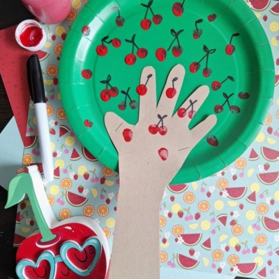 A green paper plate decorated with red fingerprints and black lines to resemble cherries, with a hand-shaped cutout attached as a tree trunk, surrounded by craft supplies and patterned paper.