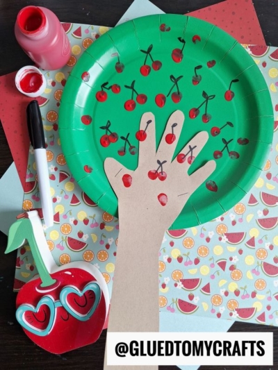 A green paper plate decorated with red fingerprints and black lines to resemble cherries, with a hand-shaped cutout attached as a tree trunk, surrounded by craft supplies and patterned paper.