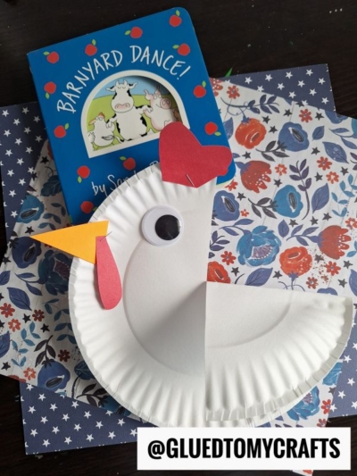 A childrens book called Barnyard Dance! sits behind a paper plate chicken craft with a googly eye, red heart comb, and yellow beak, all on floral and star-patterned paper. @GLUEDTOMYCRAFTS is written at the bottom.
