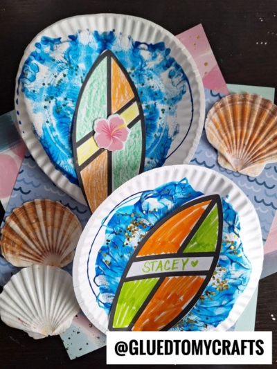 Two paper plates decorated with paint splat beach waves and colorful surfboard cutouts sit on patterned paper, surrounded by seashells. The image has a @GLUEDTOMYCRAFTS watermark in the corner.
