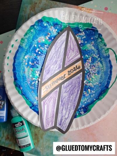 Paper plate decorated with blue and green paint splats, topped with a cutout surfboard labeled Summer 2026. Loose glitter and paint bottles are visible nearby, adding to the beach wave vibe. Handle @GLUEDTOMYCRAFTS is shown in two places.