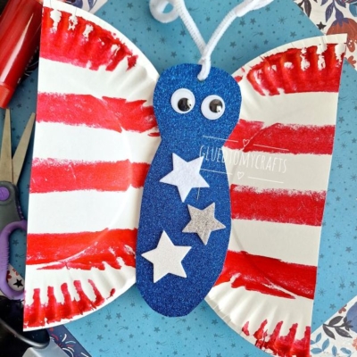 A butterfly craft made from a folded paper plate with red stripes, blue glittery body, googly eyes, white pipe cleaner antennae, and white stars, placed on patterned paper with craft supplies nearby.