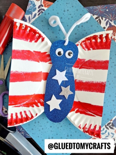 A butterfly craft made from a folded paper plate with red stripes, blue glittery body, googly eyes, white pipe cleaner antennae, and white stars, placed on patterned paper with craft supplies nearby.