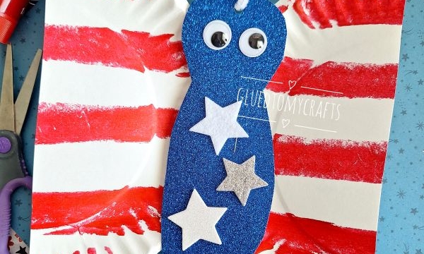 A butterfly craft made from a folded paper plate with red stripes, blue glittery body, googly eyes, white pipe cleaner antennae, and white stars, placed on patterned paper with craft supplies nearby.